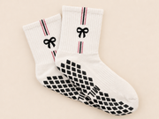 A pair of white crew socks with black bow and diamond patterns, featuring thin red and black stripes near the cuffs, displayed on a light beige background.