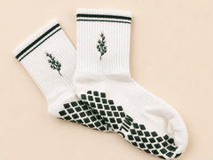 A pair of white crew socks with dark green accents, featuring a small embroidered leaf design on the upper cuff and a dark green textured grip pattern on the sole, displayed on a solid light beige background.