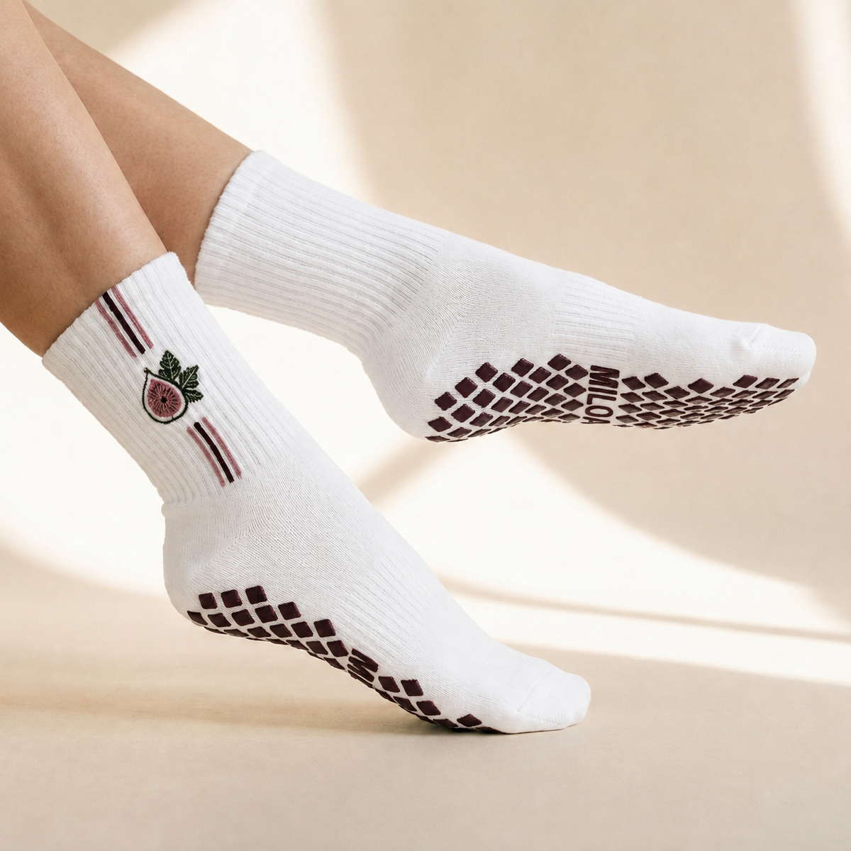 White crew socks worn on feet, featuring grip dots on the soles and a small embroidered fruit graphic with green leaves on the ankle.