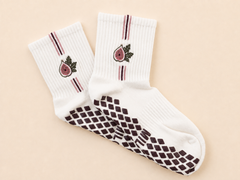 A pair of white crew socks with a dark brown diamond-patterned grip sole, featuring a pink fig illustration and thin dark stripes near the ribbed cuffs, displayed on a light beige background.
