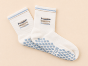 A pair of white crew socks with light blue grip patterns on the soles and light blue striped cuffs, featuring embroidered fish designs on the upper cuffs. The socks are displayed on a soft beige background.