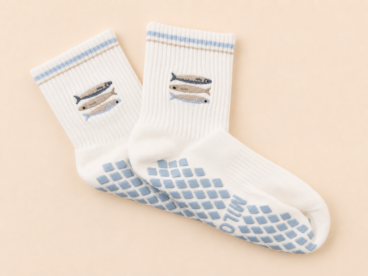 A pair of white crew socks with light blue grip patterns on the soles and light blue striped cuffs, featuring embroidered fish designs on the upper cuffs. The socks are displayed on a soft beige background.