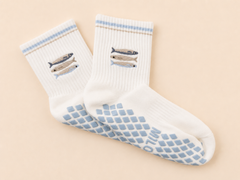 A pair of white crew socks with light blue grip patterns on the soles and light blue striped cuffs, featuring embroidered fish designs on the upper cuffs. The socks are displayed on a soft beige background.